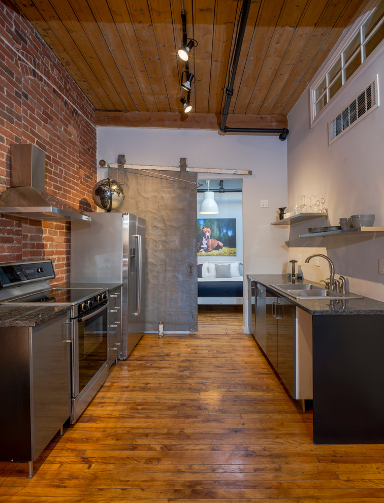 Design ideas for an industrial galley kitchen in Boston with a double-bowl sink, flat-panel cabinets, black cabinets, brick splashback, stainless steel appliances, medium hardwood floors, no island and black benchtop.