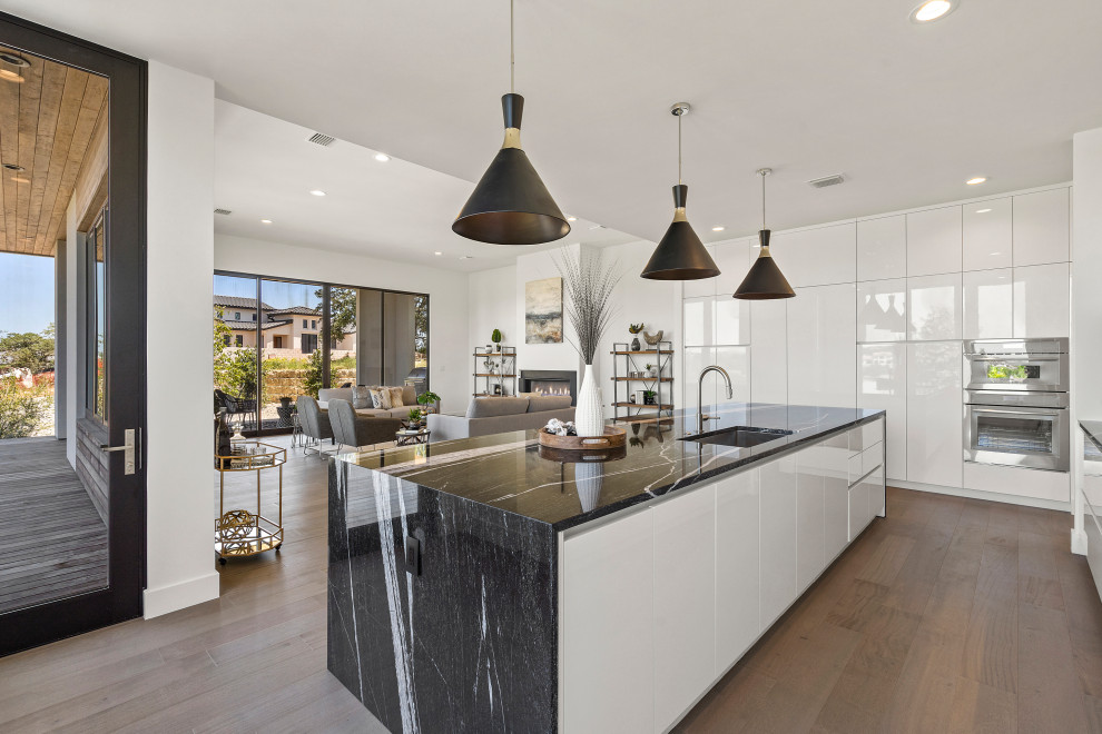 Mid-sized contemporary galley eat-in kitchen in Austin with an undermount sink, flat-panel cabinets, white cabinets, granite benchtops, black splashback, granite splashback, stainless steel appliances, medium hardwood floors, with island, brown floor and black benchtop.