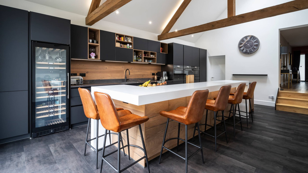 Mid-sized contemporary galley open plan kitchen in Oxfordshire with flat-panel cabinets, black cabinets, quartzite benchtops, brown splashback, timber splashback, black appliances, with island, black floor, multi-coloured benchtop, exposed beam and vaulted.