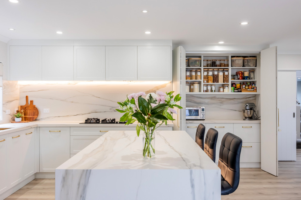Inspiration for a mid-sized modern l-shaped open plan kitchen in Auckland with a double-bowl sink, shaker cabinets, white cabinets, quartz benchtops, white splashback, engineered quartz splashback, black appliances, light hardwood floors, with island, grey floor and white benchtop.
