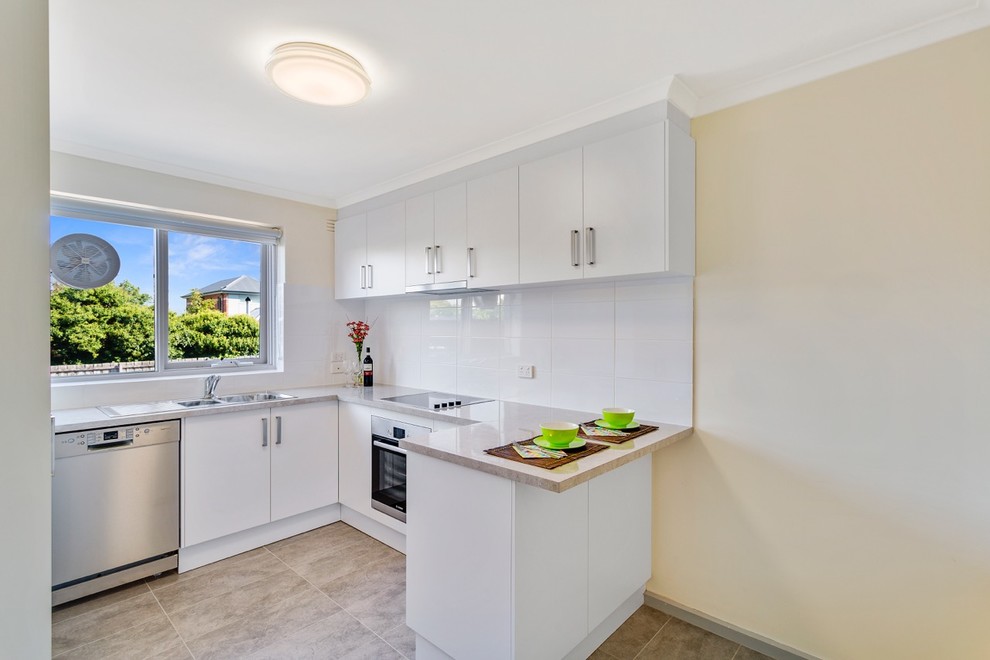This is an example of a small contemporary u-shaped eat-in kitchen in Melbourne with a double-bowl sink, flat-panel cabinets, white cabinets, marble benchtops, white splashback, porcelain splashback, stainless steel appliances, travertine floors, a peninsula and brown floor.