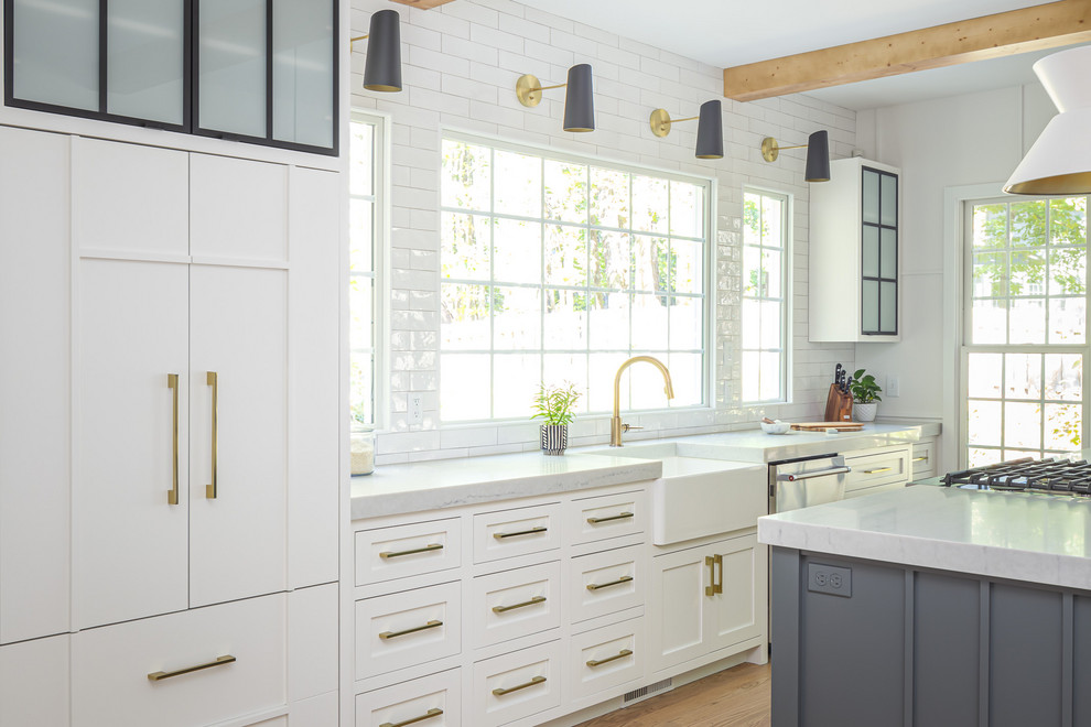 Example of a mid-sized farmhouse single-wall light wood floor open concept kitchen design in Raleigh with a farmhouse sink, flat-panel cabinets, white cabinets, quartzite countertops, white backsplash, subway tile backsplash, paneled appliances, an island and white countertops