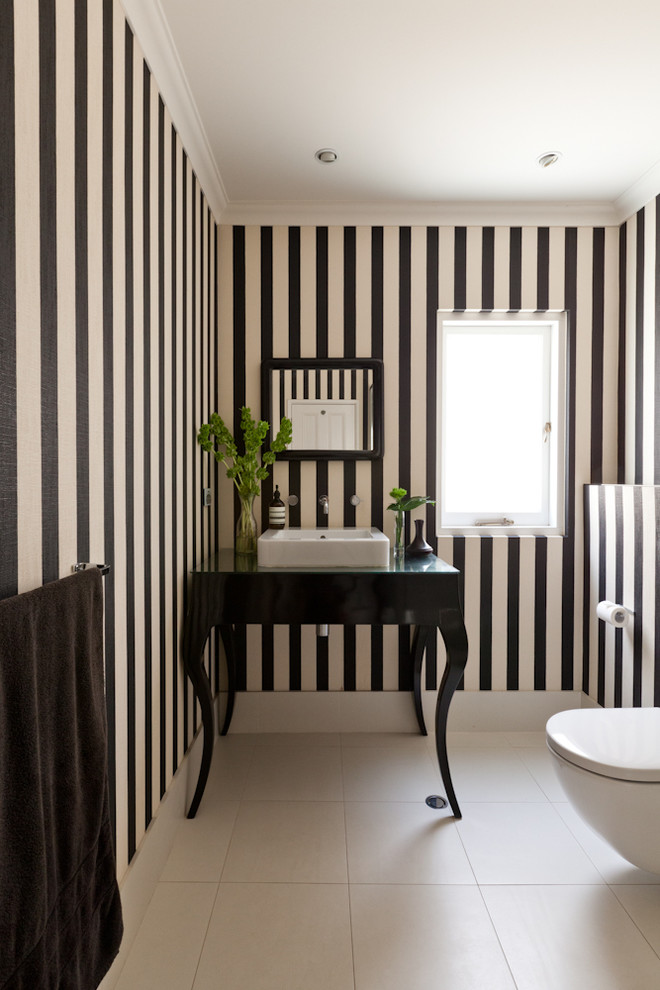 This is an example of a mid-sized transitional powder room in Perth with a vessel sink, furniture-like cabinets, black cabinets, glass benchtops, a wall-mount toilet, beige tile, multi-coloured walls and ceramic floors.