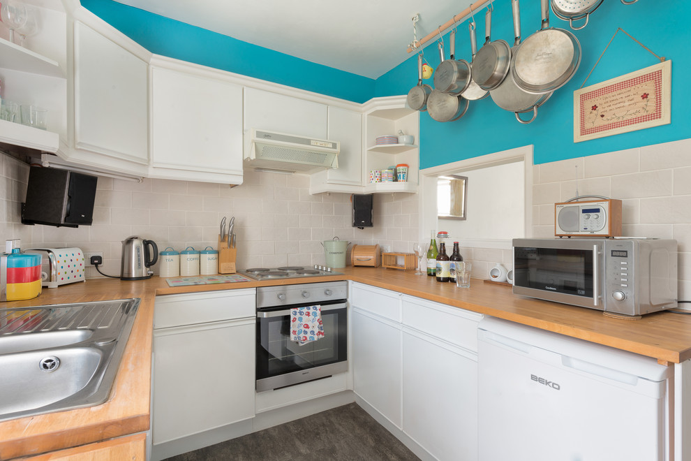 Design ideas for a small eclectic u-shaped separate kitchen in Devon with a drop-in sink, flat-panel cabinets, white cabinets, wood benchtops, white splashback, ceramic splashback, stainless steel appliances and linoleum floors.