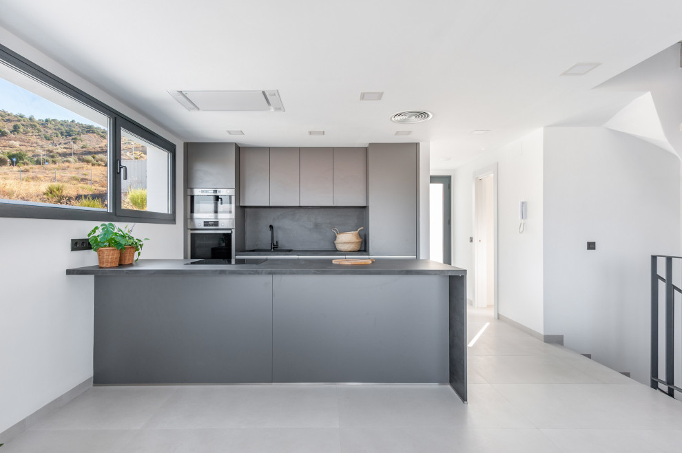 Mid-sized modern galley open plan kitchen in Malaga with a drop-in sink, recessed-panel cabinets, quartz benchtops, black splashback, engineered quartz splashback, stainless steel appliances, ceramic floors, with island, grey floor and black benchtop.