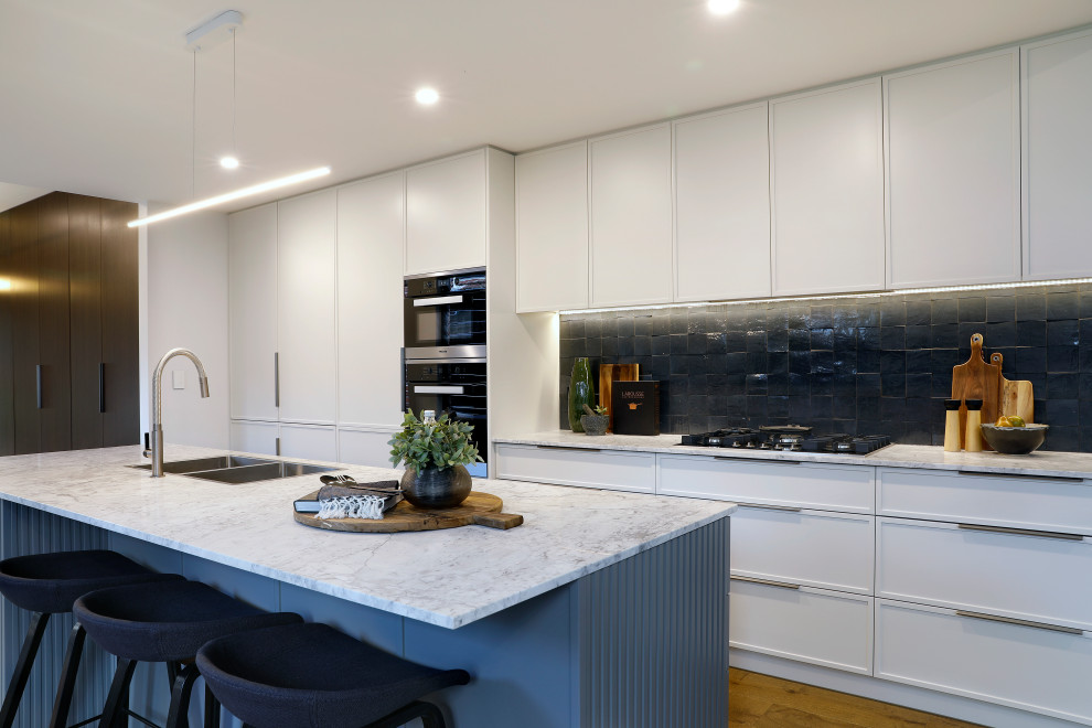 Mid-sized contemporary galley open plan kitchen in Sydney with a drop-in sink, marble benchtops, ceramic splashback, stainless steel appliances, with island, white benchtop, flat-panel cabinets, white cabinets, black splashback, medium hardwood floors and brown floor.