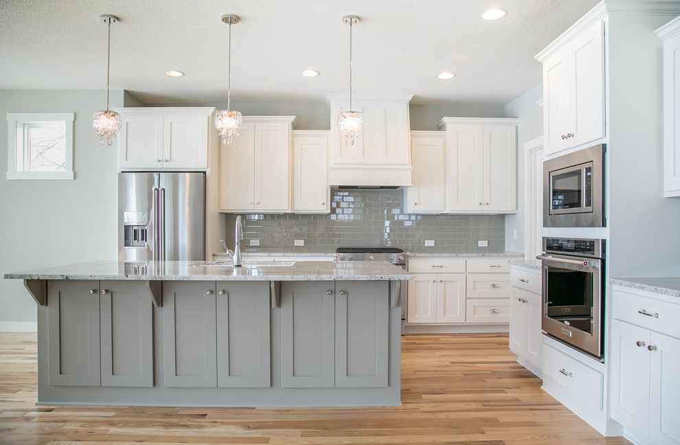 Photo of a transitional l-shaped eat-in kitchen in Minneapolis with a drop-in sink, shaker cabinets, white cabinets, granite benchtops, grey splashback, ceramic splashback, stainless steel appliances, light hardwood floors and with island.