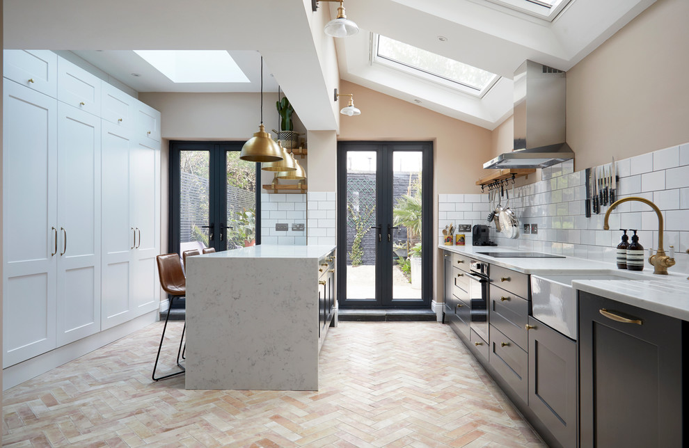 This is an example of a mid-sized transitional single-wall kitchen in London with a farmhouse sink, grey cabinets, solid surface benchtops, white splashback, ceramic splashback, stainless steel appliances, ceramic floors, with island, white benchtop, shaker cabinets and pink floor.