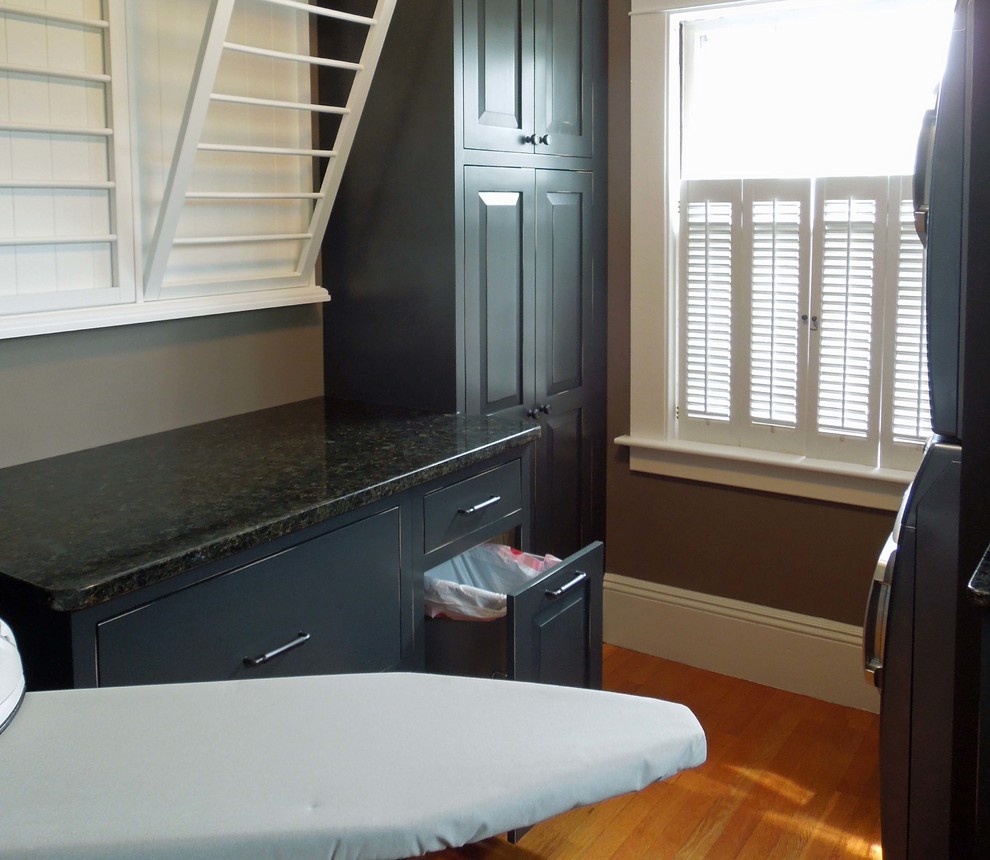 This is an example of a mid-sized transitional galley dedicated laundry room in Charleston with raised-panel cabinets, distressed cabinets, granite benchtops, medium hardwood floors and a stacked washer and dryer.