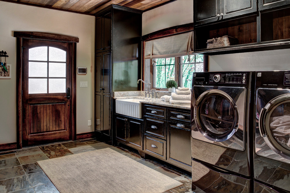 Mid-sized country single-wall utility room in Minneapolis with a farmhouse sink, raised-panel cabinets, black cabinets, granite benchtops, beige walls, slate floors and a side-by-side washer and dryer.
