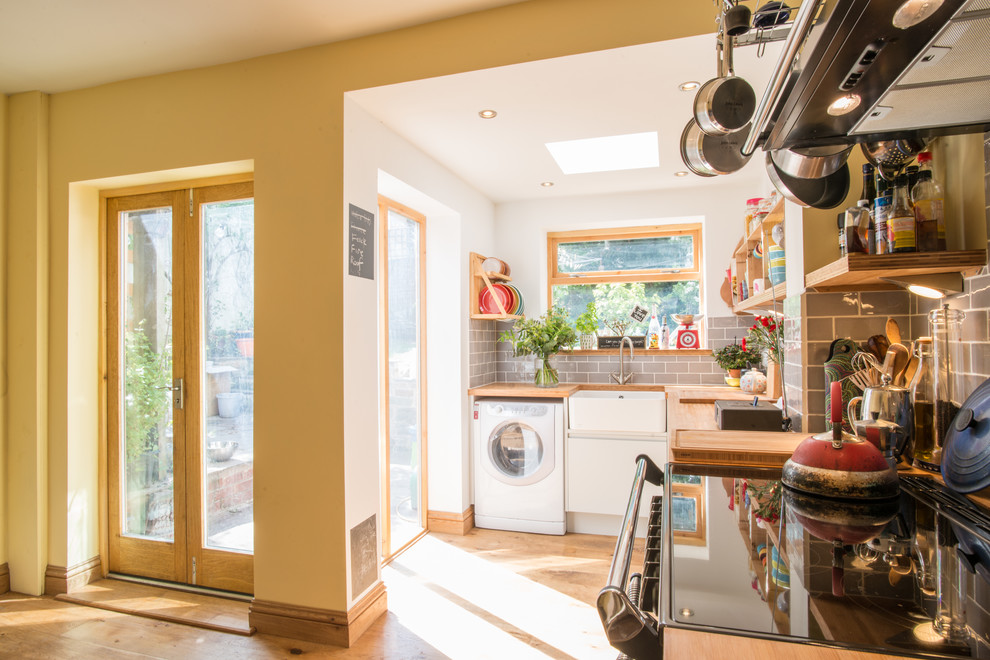 This is an example of a mid-sized eclectic l-shaped eat-in kitchen in Other with a farmhouse sink, flat-panel cabinets, grey cabinets, wood benchtops, grey splashback, subway tile splashback, white appliances, medium hardwood floors and no island.
