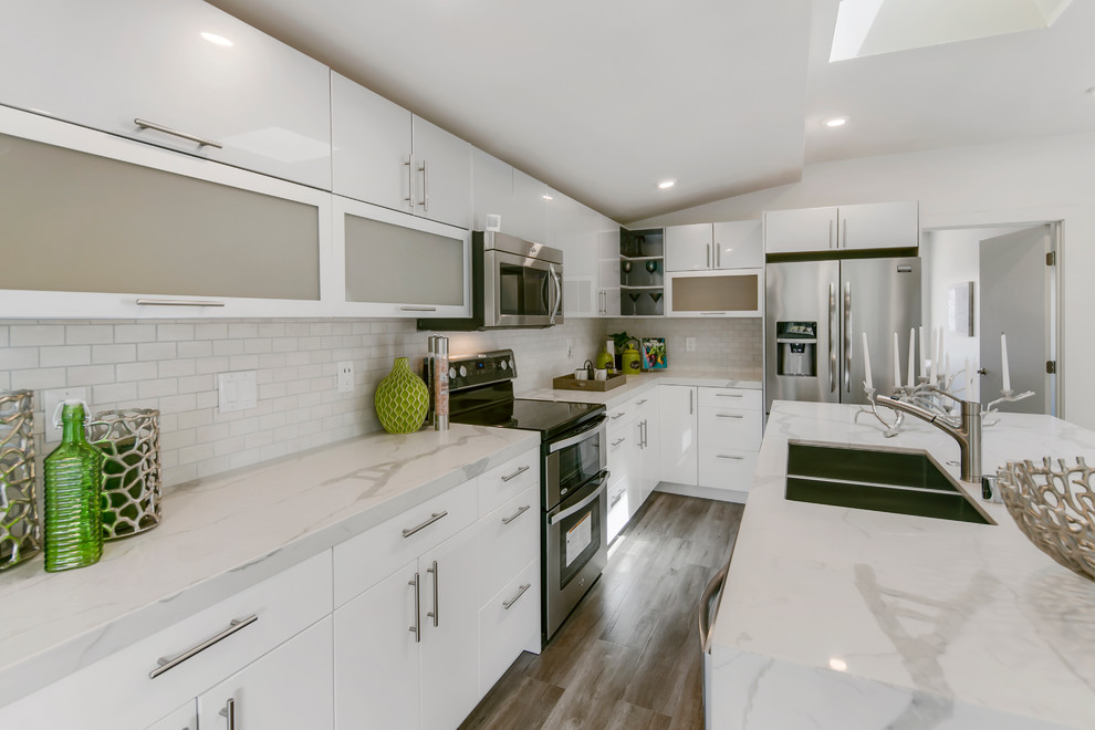 Photo of a mid-sized modern l-shaped open plan kitchen in San Francisco with a double-bowl sink, flat-panel cabinets, white cabinets, marble benchtops, white splashback, ceramic splashback, stainless steel appliances, with island and laminate floors.