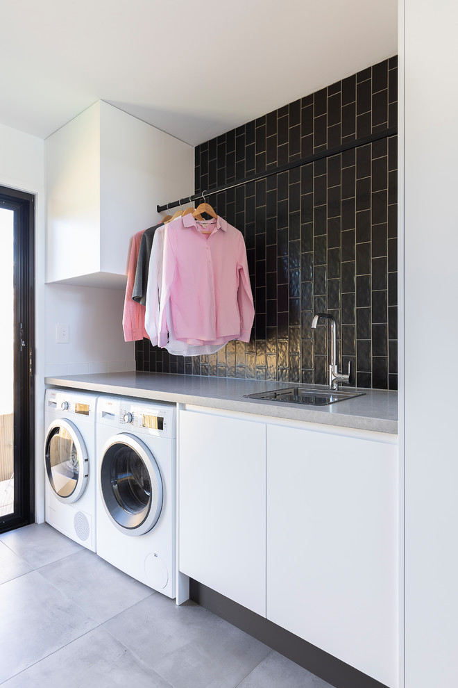 Photo of a mid-sized contemporary single-wall utility room in Brisbane with a single-bowl sink, flat-panel cabinets, white cabinets, quartz benchtops, black walls, ceramic floors, a side-by-side washer and dryer, grey floor and grey benchtop.