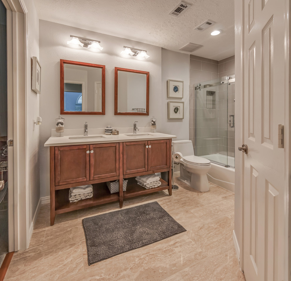 Design ideas for a mid-sized mediterranean 3/4 bathroom in Houston with beaded inset cabinets, medium wood cabinets, an alcove tub, a shower/bathtub combo, a two-piece toilet, gray tile, subway tile, white walls, an undermount sink, solid surface benchtops, beige floor and a sliding shower screen.
