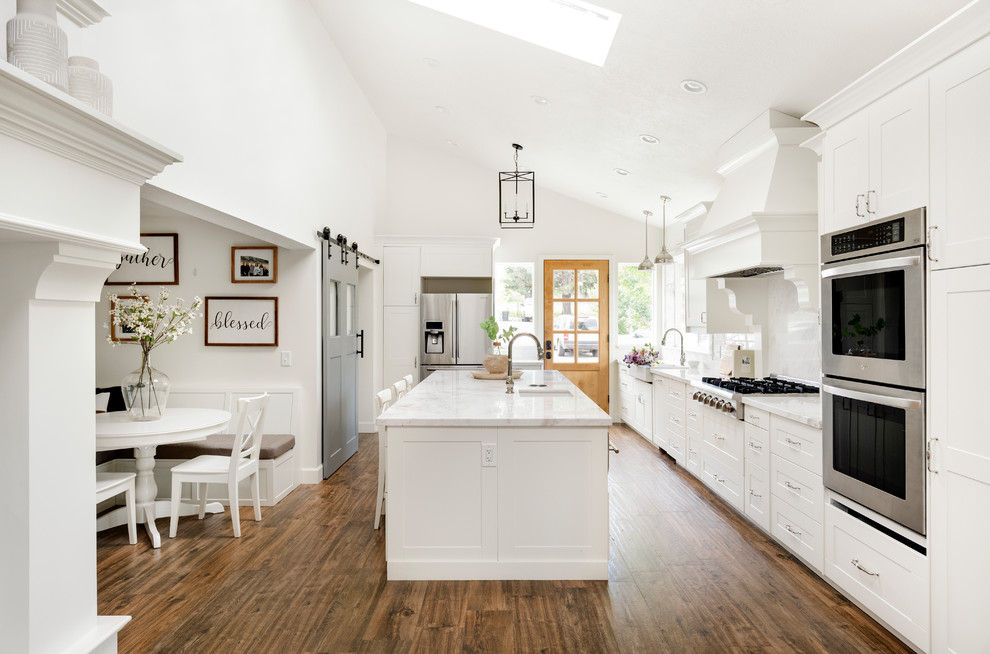 Photo of a mid-sized country galley eat-in kitchen in Salt Lake City with a farmhouse sink, shaker cabinets, white cabinets, marble benchtops, white splashback, marble splashback, stainless steel appliances, porcelain floors, with island, brown floor and white benchtop.