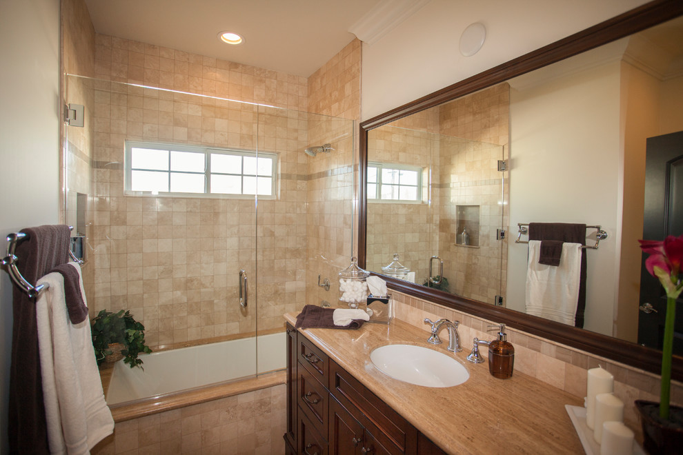 This is an example of a mid-sized mediterranean bathroom in Los Angeles with recessed-panel cabinets, dark wood cabinets, an undermount tub, a shower/bathtub combo, a one-piece toilet, beige tile, travertine, beige walls, travertine floors, an undermount sink, limestone benchtops and a sliding shower screen.