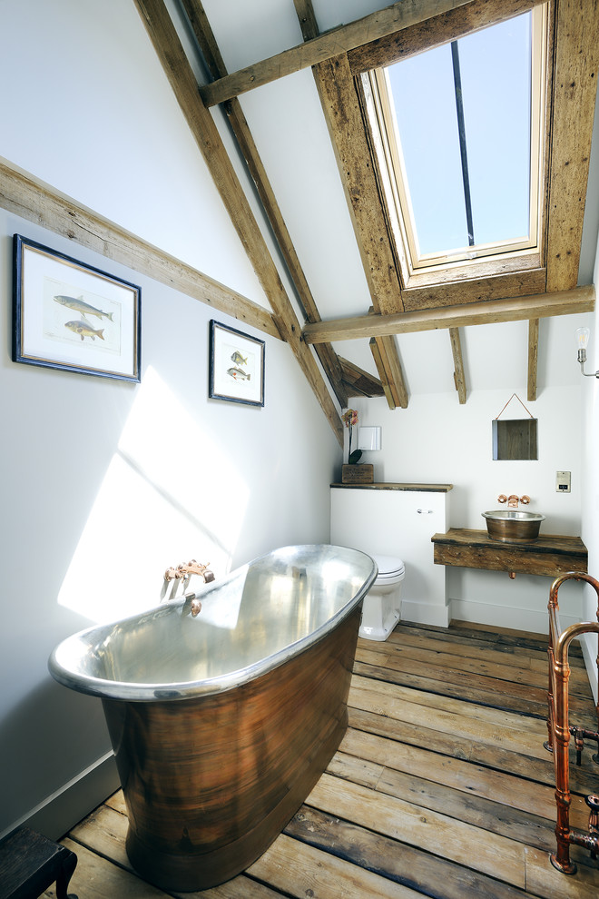This is an example of a country bathroom in Hampshire with a vessel sink, wood benchtops, a freestanding tub, a two-piece toilet, white walls, light hardwood floors and brown benchtops.