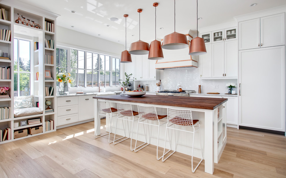 Photo of a country l-shaped kitchen in Calgary with a farmhouse sink, shaker cabinets, white cabinets, wood benchtops, white splashback, panelled appliances, light hardwood floors, with island, beige floor and brown benchtop.