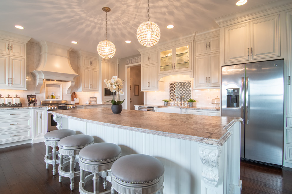 Traditional u-shaped kitchen in Philadelphia with a farmhouse sink, raised-panel cabinets, white cabinets, white splashback, stainless steel appliances, dark hardwood floors, with island, brown floor and grey benchtop.