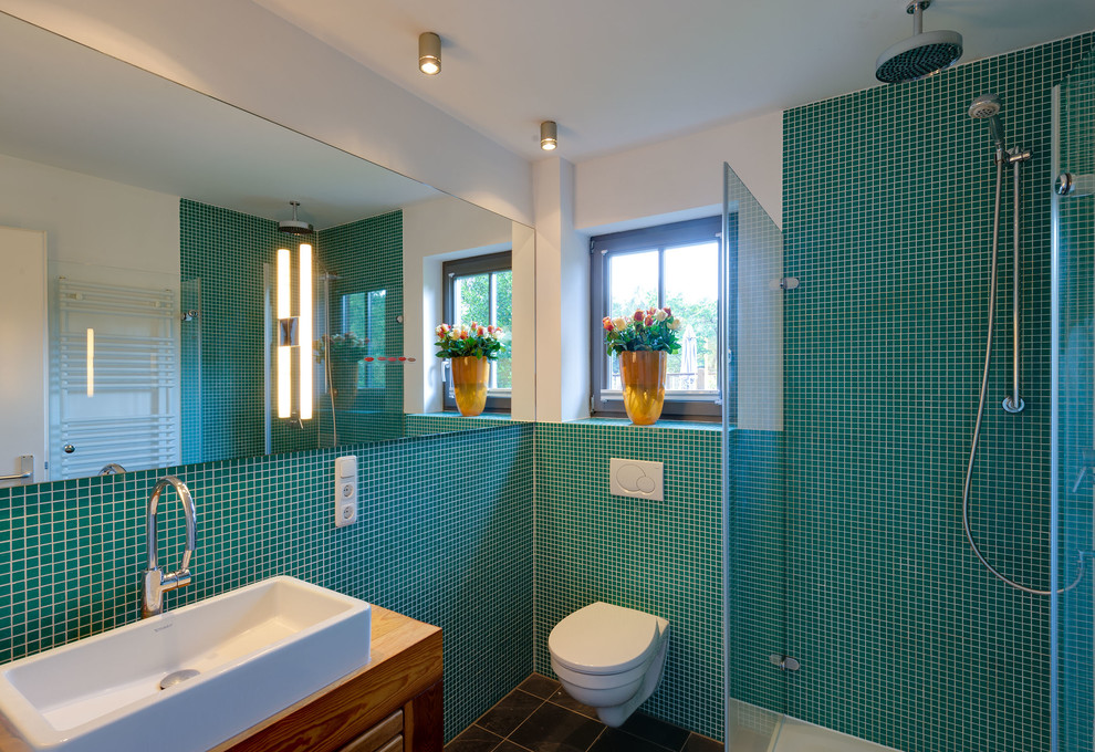 This is an example of a small contemporary bathroom in Hamburg with medium wood cabinets, a wall-mount toilet, blue tile, mosaic tile, white walls, a vessel sink and wood benchtops.