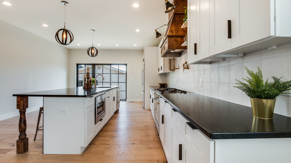 Example of a mid-sized country galley light wood floor and beige floor eat-in kitchen design in Denver with an undermount sink, shaker cabinets, white cabinets, white backsplash, ceramic backsplash, stainless steel appliances, an island and black countertops