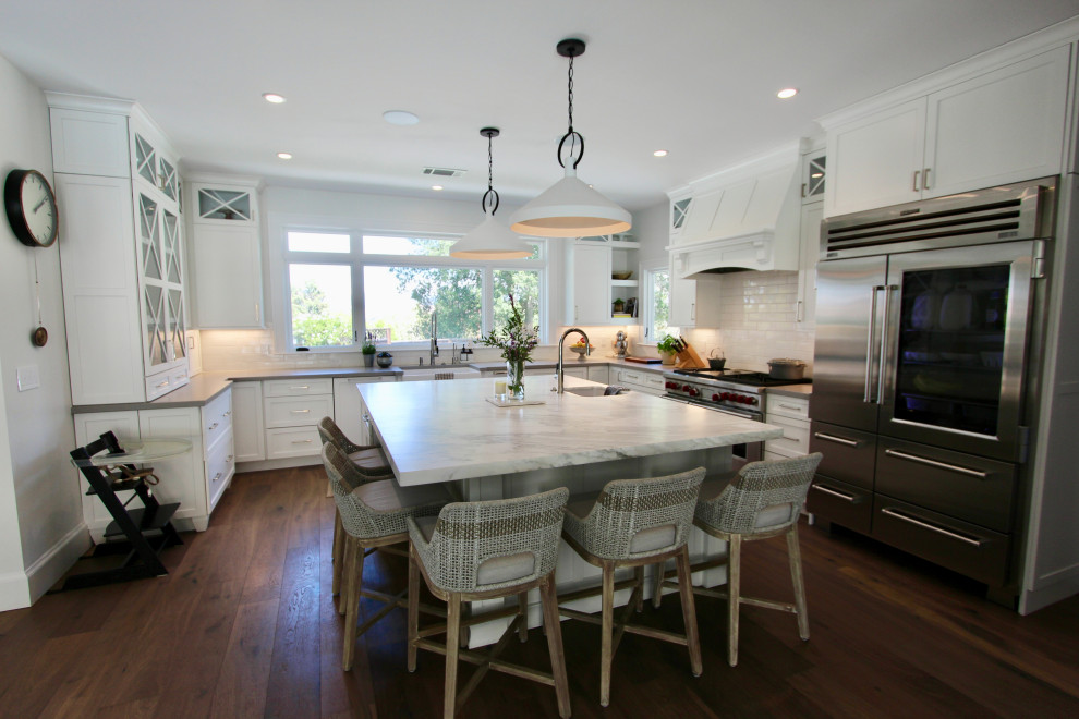 Photo of a large farmhouse u-shaped open plan kitchen in San Francisco with a belfast sink, recessed-panel cabinets, white cabinets, marble worktops, white splashback, metro tiled splashback, stainless steel appliances, medium hardwood flooring, an island, white worktops and feature lighting.