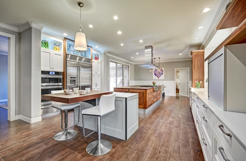 This is an example of a large transitional galley eat-in kitchen in Sacramento with a farmhouse sink, recessed-panel cabinets, white cabinets, quartz benchtops, stainless steel appliances, medium hardwood floors, multiple islands, white splashback and porcelain splashback.