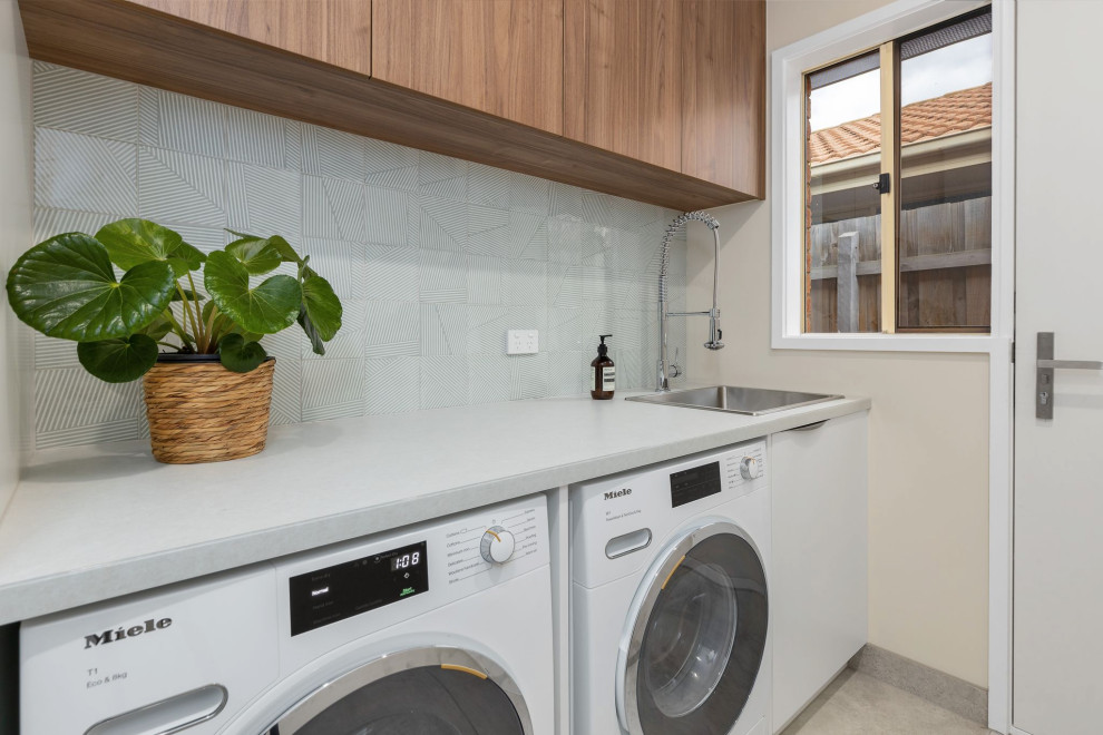 This is an example of a mid-sized contemporary single-wall dedicated laundry room in Melbourne with flat-panel cabinets, light wood cabinets, grey walls, porcelain floors, grey floor, a drop-in sink, quartz benchtops, grey splashback, porcelain splashback, a side-by-side washer and dryer and white benchtop.