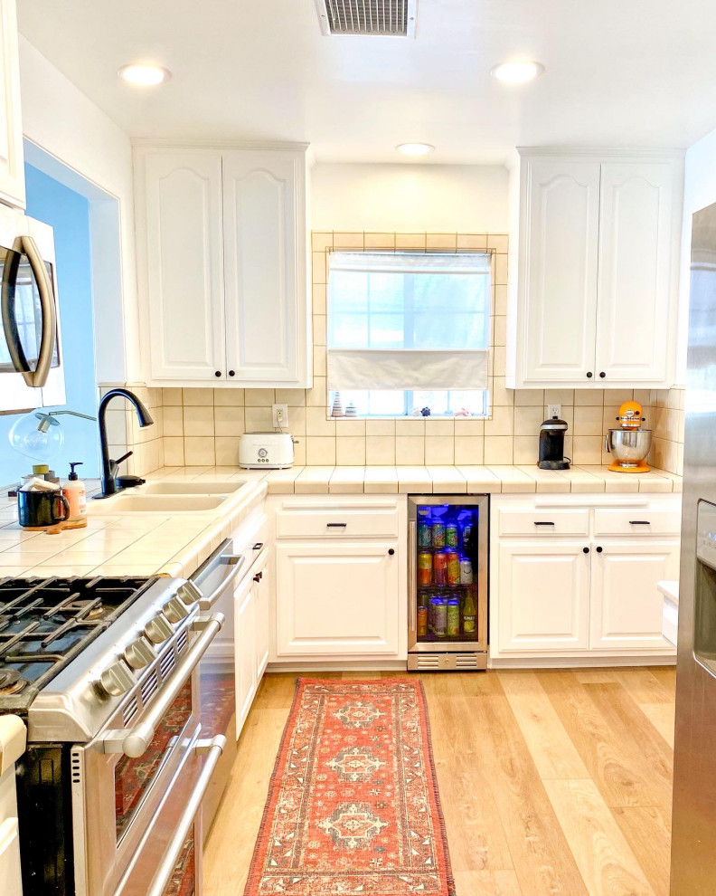 This is an example of a medium sized traditional l-shaped kitchen pantry in Orange County with a double-bowl sink, white cabinets, tile countertops, beige splashback, ceramic splashback, vinyl flooring and beige floors.