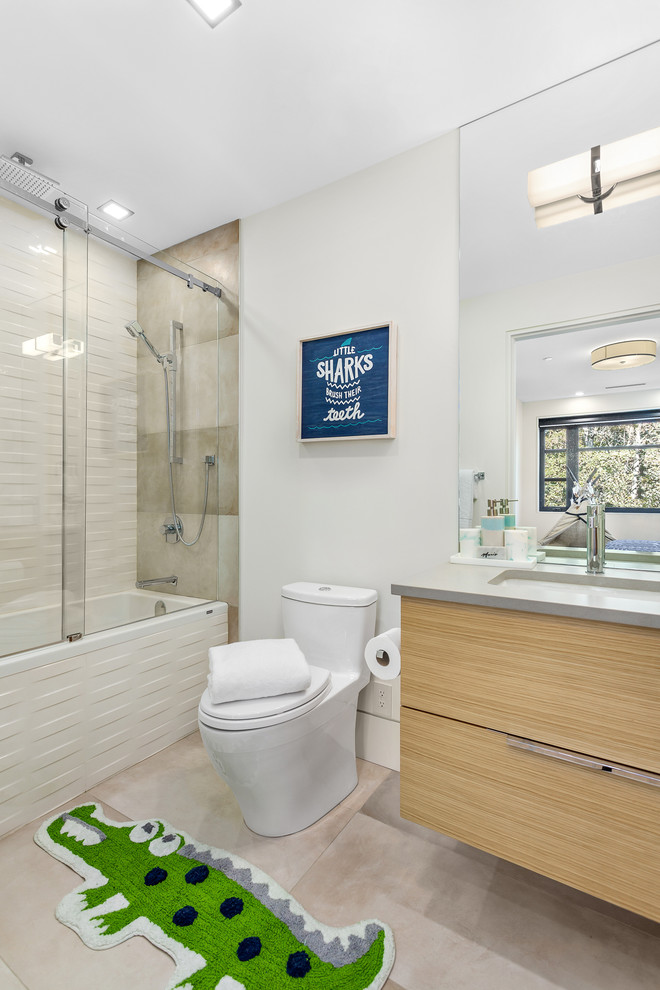 Photo of a mid-sized contemporary kids bathroom in Vancouver with flat-panel cabinets, light wood cabinets, a drop-in tub, a shower/bathtub combo, a two-piece toilet, gray tile, porcelain tile, grey walls, porcelain floors, an undermount sink, engineered quartz benchtops, grey floor, a sliding shower screen and grey benchtops.