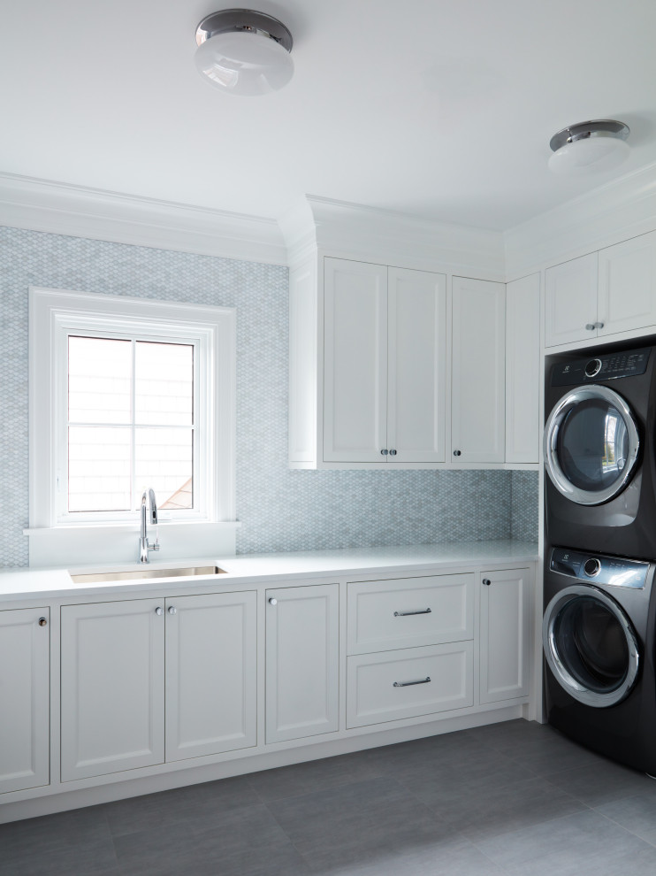 Inredning av en klassisk stor vita l-formad vitt tvättstuga enbart för tvätt, med en undermonterad diskho, luckor med infälld panel, vita skåp, blått stänkskydd, stänkskydd i mosaik, flerfärgade väggar, klinkergolv i porslin, en tvättpelare och grått golv