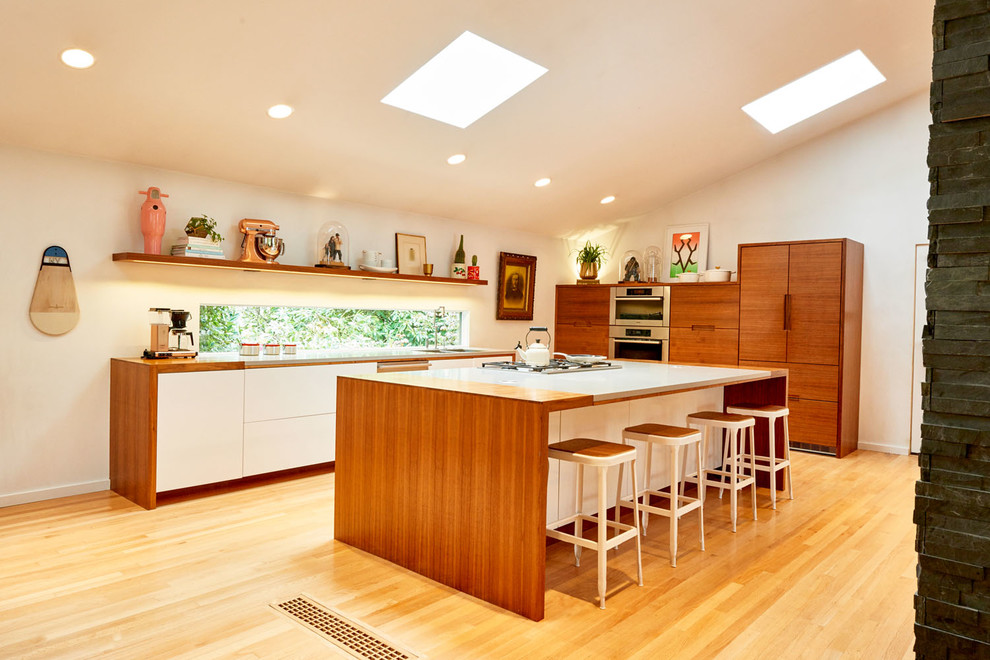 This is an example of a large midcentury l-shaped open plan kitchen in Portland with an undermount sink, flat-panel cabinets, medium wood cabinets, solid surface benchtops, panelled appliances, light hardwood floors, with island, beige floor and white benchtop.