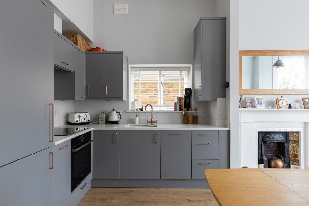 Small arts and crafts l-shaped open plan kitchen in London with an integrated sink, flat-panel cabinets, grey cabinets, solid surface benchtops, white splashback, limestone splashback, stainless steel appliances, laminate floors and no island.