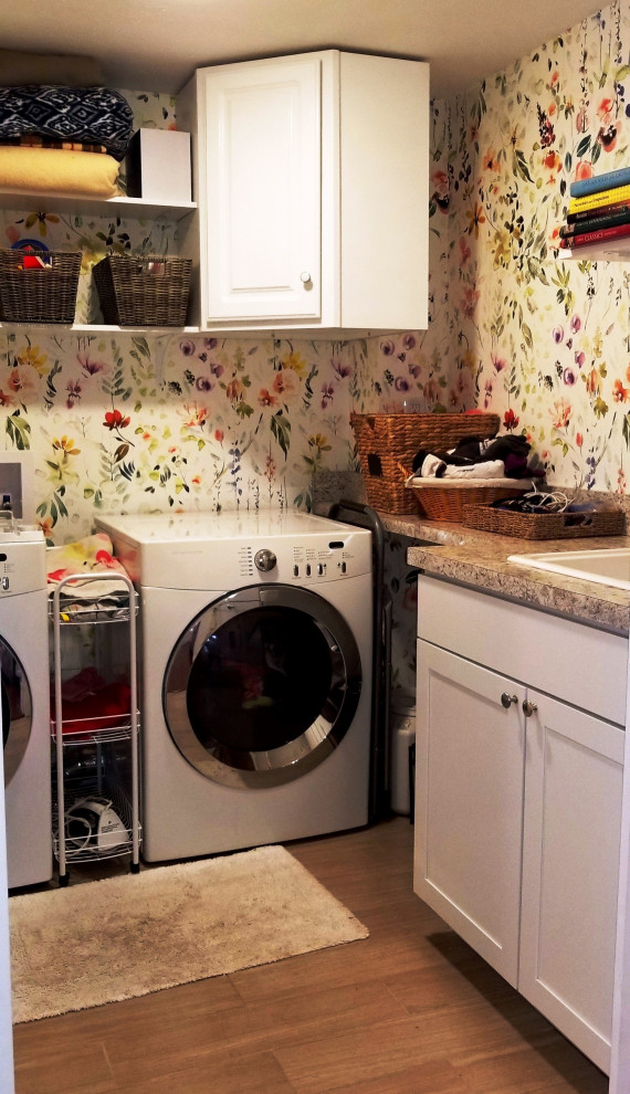 Immagine di una sala lavanderia tradizionale di medie dimensioni con lavello da incasso, ante bianche, top in laminato, pareti multicolore, pavimento in laminato, lavatrice e asciugatrice affiancate, pavimento marrone, top multicolore e carta da parati