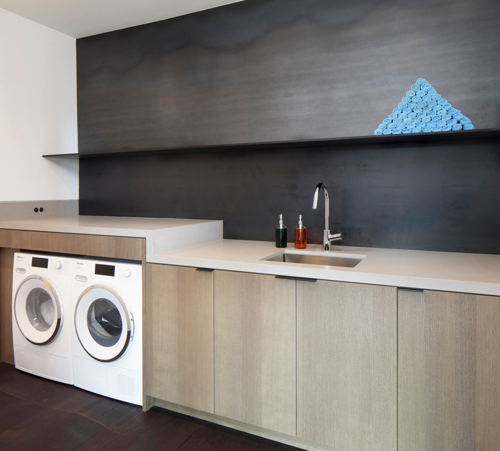 This is an example of a mid-sized modern galley dedicated laundry room in San Francisco with an undermount sink, flat-panel cabinets, medium wood cabinets, quartz benchtops, grey splashback, metal splashback, white walls, dark hardwood floors, a side-by-side washer and dryer, grey floor and white benchtop.
