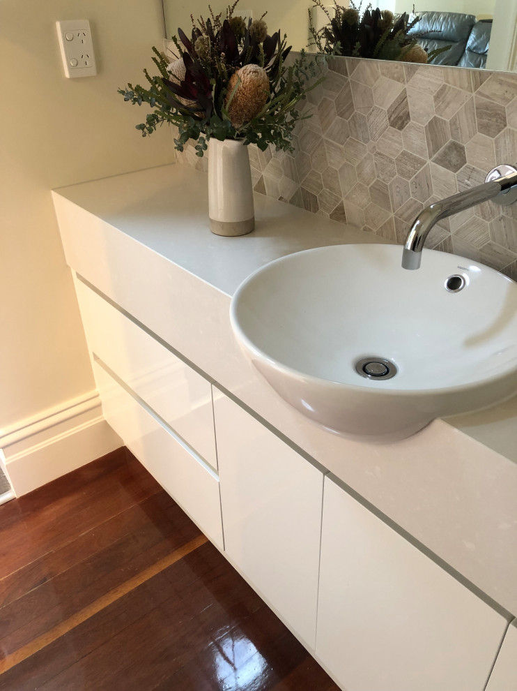 This is an example of a mid-sized contemporary powder room in Other with furniture-like cabinets, white cabinets, multi-coloured tile, ceramic tile, multi-coloured walls, medium hardwood floors, an integrated sink, quartzite benchtops, brown floor and beige benchtops.
