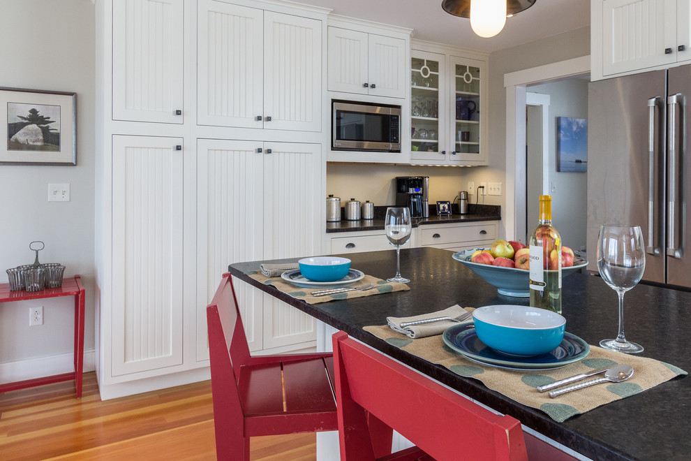 Beach style kitchen in Portland Maine with a farmhouse sink, beaded inset cabinets, white cabinets, granite benchtops, black splashback, stainless steel appliances, medium hardwood floors and with island.