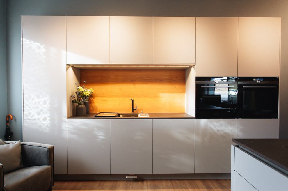 This is an example of a large contemporary galley eat-in kitchen in Other with an undermount sink, flat-panel cabinets, grey cabinets, granite benchtops, brown splashback, timber splashback, black appliances, bamboo floors, with island, brown floor and black benchtop.