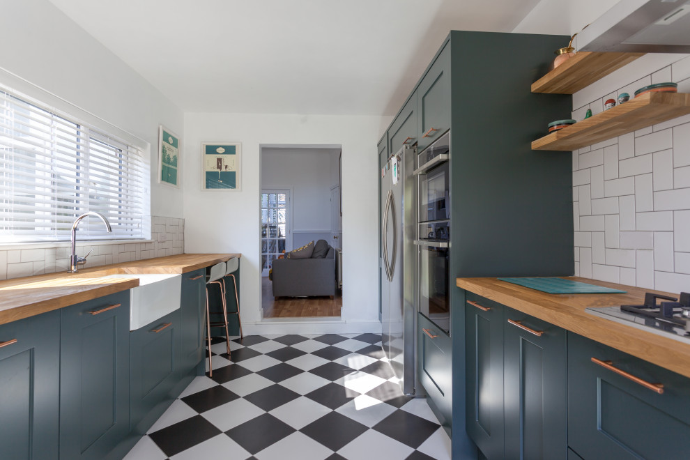 This is an example of a mid-sized transitional galley separate kitchen in Manchester with a farmhouse sink, shaker cabinets, green cabinets, wood benchtops, white splashback, subway tile splashback, stainless steel appliances, porcelain floors, a peninsula, white floor and brown benchtop.