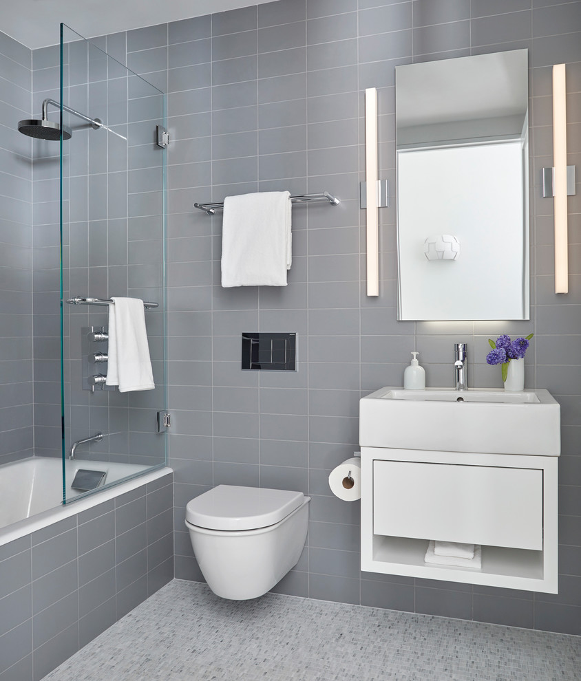 This is an example of a mid-sized contemporary master bathroom in New York with flat-panel cabinets, white cabinets, an alcove tub, a shower/bathtub combo, a wall-mount toilet, gray tile, porcelain tile, grey walls, mosaic tile floors, a vessel sink, grey floor and a hinged shower door.