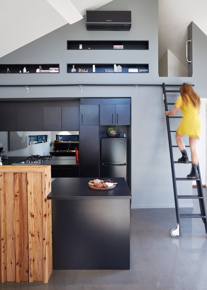 This is an example of a contemporary open plan kitchen in Melbourne with flat-panel cabinets, black cabinets, metallic splashback, mirror splashback, black appliances, concrete floors and with island.