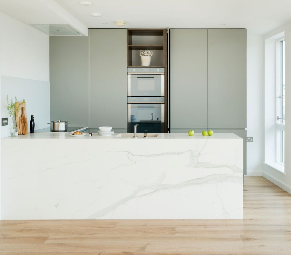 Design ideas for a mid-sized contemporary u-shaped kitchen in London with flat-panel cabinets, glass sheet splashback, light hardwood floors, an undermount sink, green cabinets, a peninsula, solid surface benchtops, grey splashback, brown floor and white benchtop.