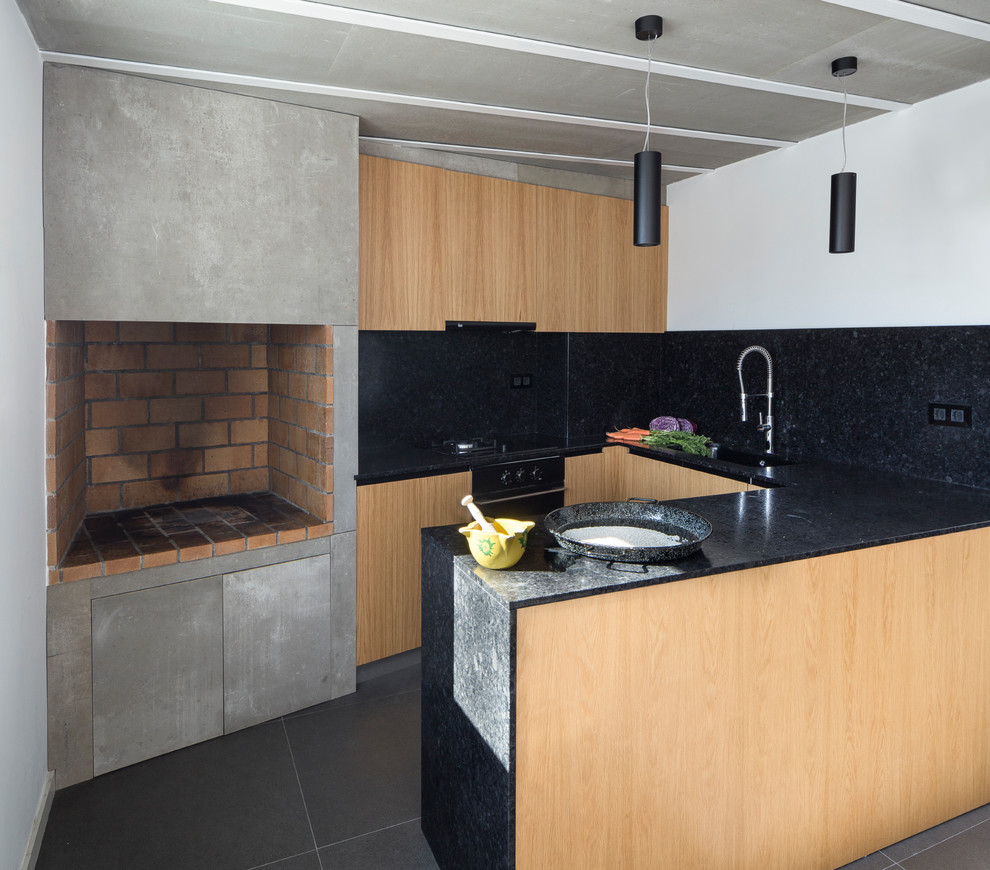 Design ideas for an industrial u-shaped eat-in kitchen in Valencia with flat-panel cabinets, black benchtop, light wood cabinets, black splashback, black appliances, a peninsula and grey floor.