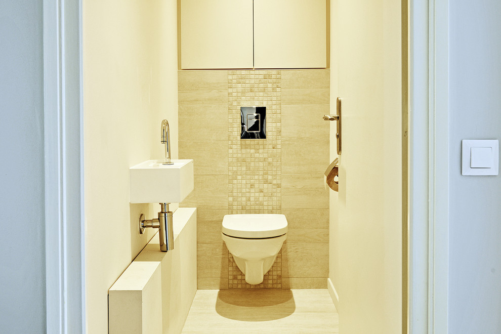 Photo of a small modern powder room in Lyon with beaded inset cabinets, beige cabinets, a wall-mount toilet, beige tile, mosaic tile, beige walls, ceramic floors, a wall-mount sink, solid surface benchtops, beige floor and white benchtops.