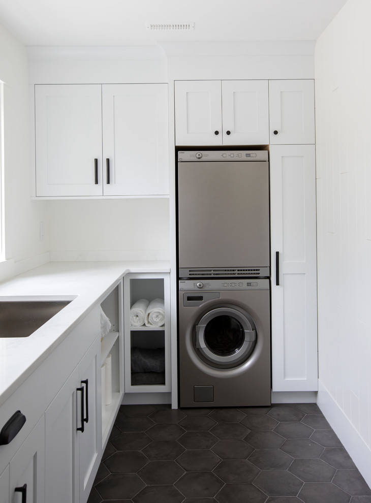 Country galley dedicated laundry room in Other with shaker cabinets, white cabinets, marble benchtops, white walls, terra-cotta floors, a stacked washer and dryer, black floor and an undermount sink.