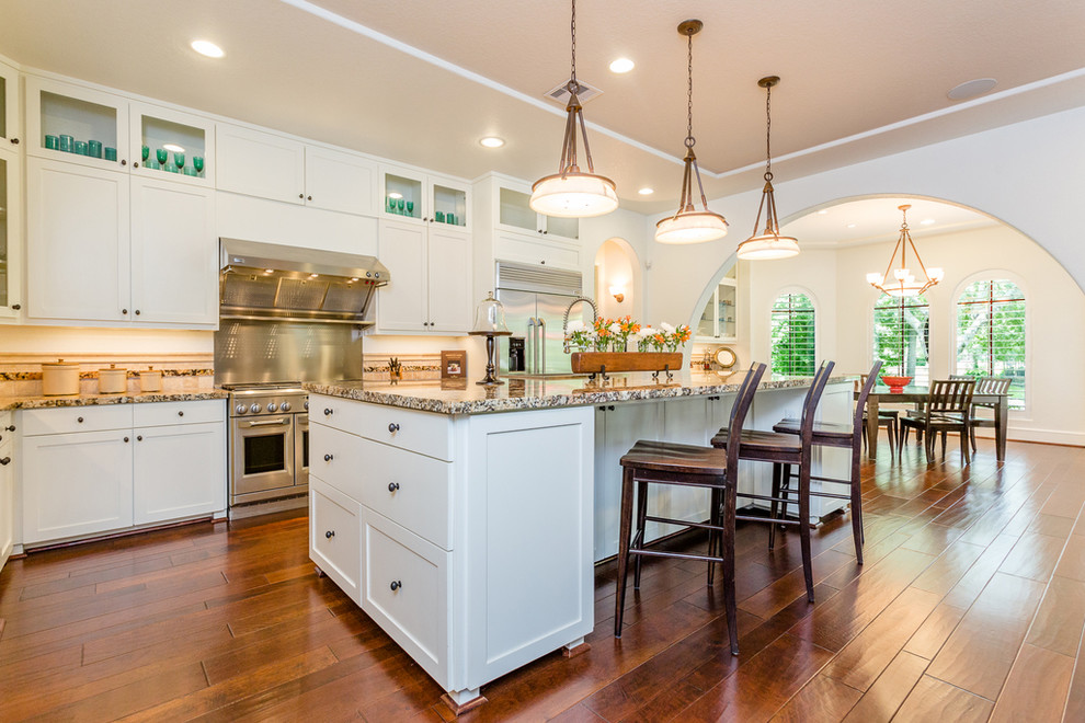 Large mediterranean l-shaped eat-in kitchen in Houston with shaker cabinets, white cabinets, granite benchtops, stainless steel appliances, medium hardwood floors, with island, multi-coloured splashback and multi-coloured benchtop.