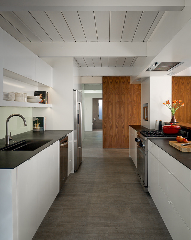 This is an example of a small midcentury galley eat-in kitchen in San Francisco with an undermount sink, flat-panel cabinets, white cabinets, white splashback, glass sheet splashback, stainless steel appliances, porcelain floors, solid surface benchtops, with island, grey floor and exposed beam.
