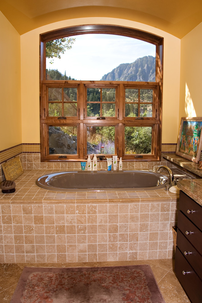 This is an example of a mid-sized country master bathroom in Denver with flat-panel cabinets, dark wood cabinets, granite benchtops, a drop-in tub, beige tile, stone tile, yellow walls and travertine floors.