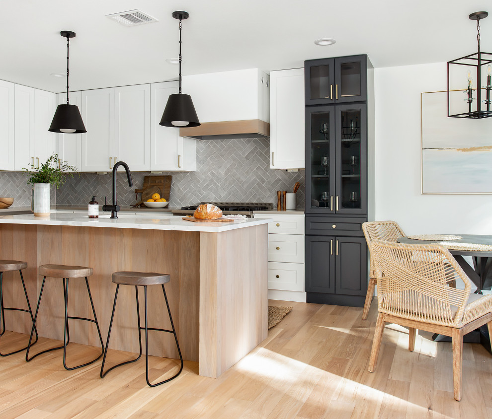 Mid-sized beach style l-shaped eat-in kitchen in Dallas with a single-bowl sink, shaker cabinets, light wood cabinets, quartz benchtops, grey splashback, porcelain splashback, stainless steel appliances, light hardwood floors, with island, brown floor and white benchtop.