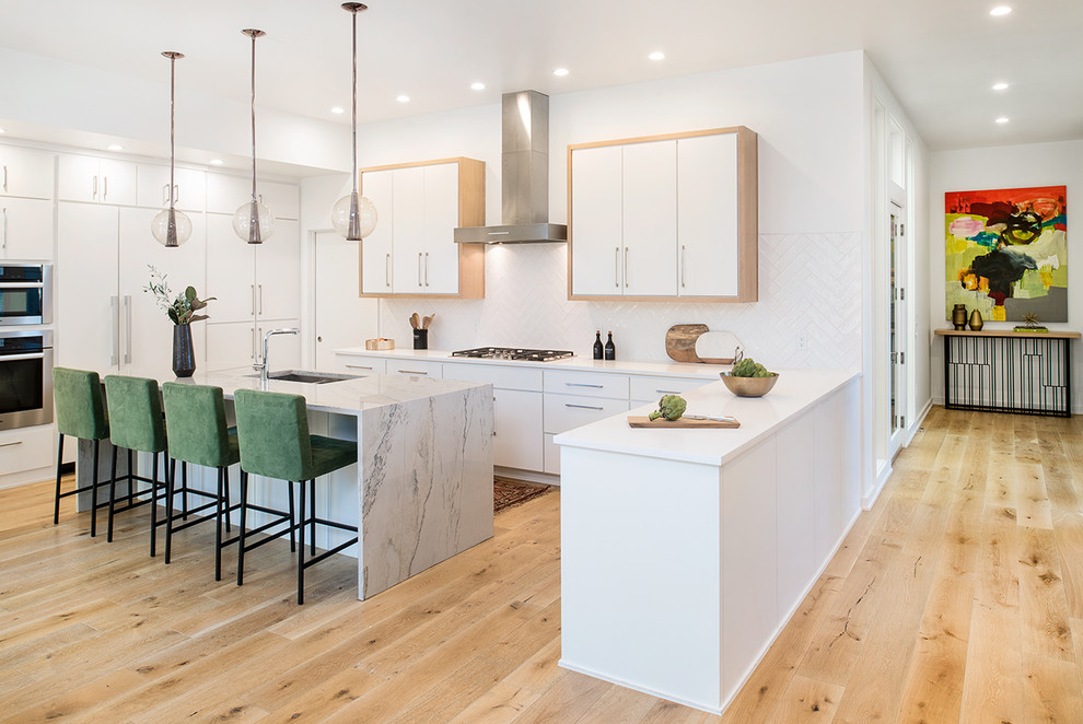 This is an example of a contemporary galley open plan kitchen in Austin with flat-panel cabinets, white cabinets, white splashback, stainless steel appliances, light hardwood floors, a peninsula and brown floor.